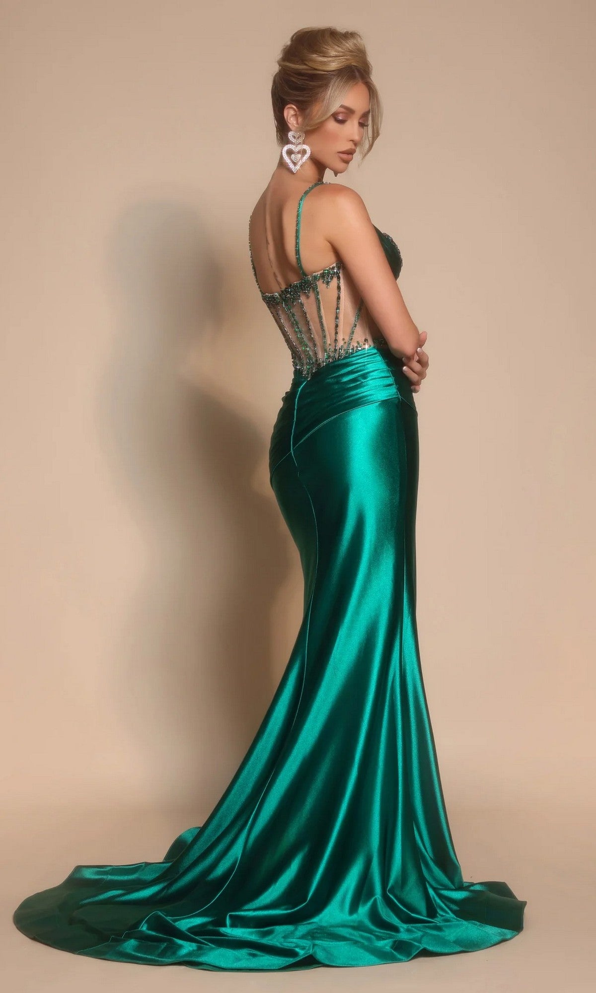 A woman stands in profile against a beige background wearing the Portia and Scarlett PS26158, a fitted emerald green satin long formal dress with a corset bodice, sheer beaded back, and train.